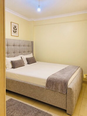 A single bed with a tufted headboard, a bedside table, and a small rug on the floor. - Stylish 1 bedroom apartment (Nairobi)