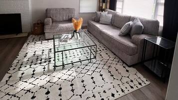 A modern living room with a glass coffee table, grey sofas, a patterned rug, and a fireplace.