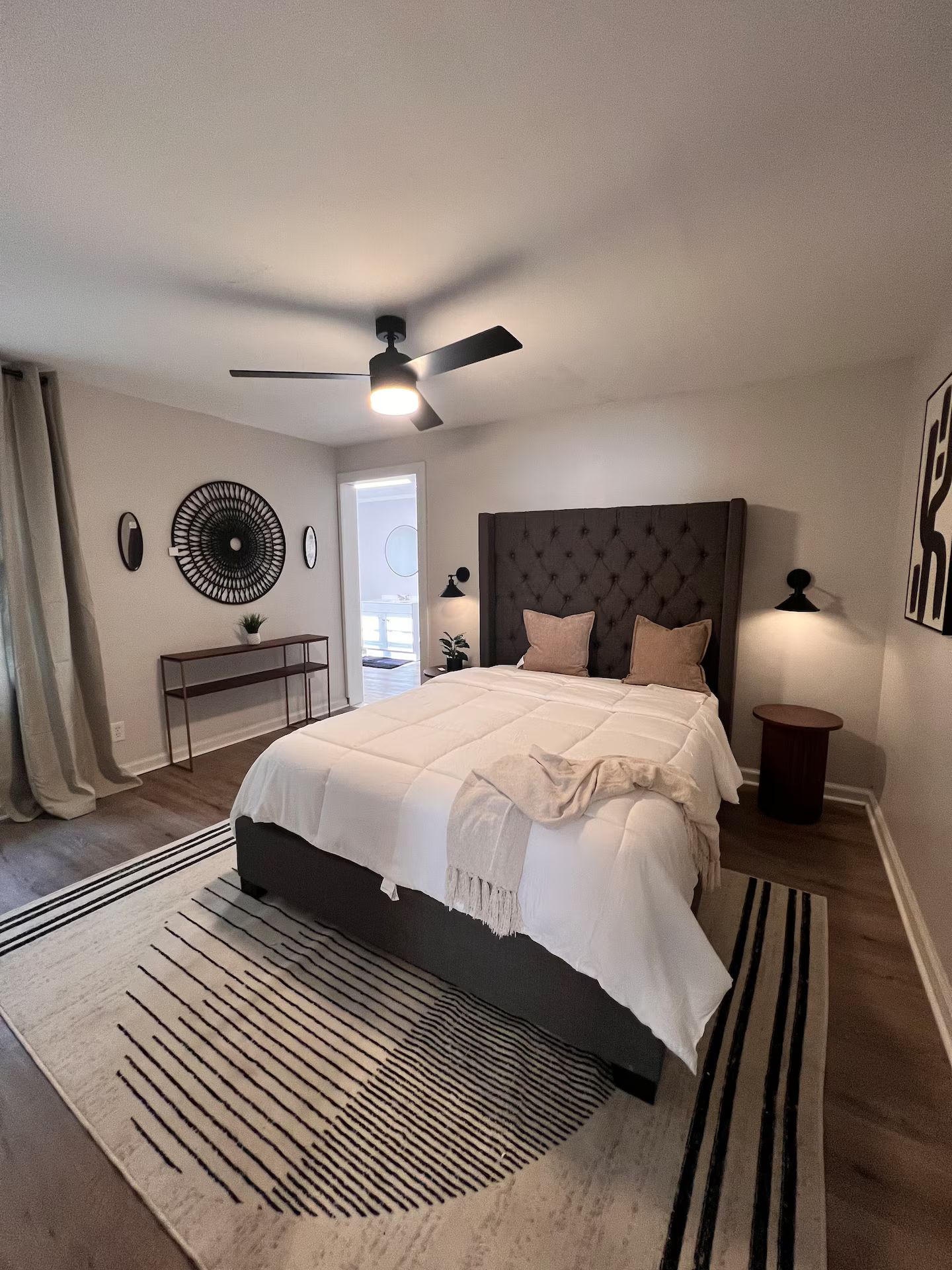 A bedroom with a bed, a ceiling fan, a nightstand, a lamp, and a view of the outside.