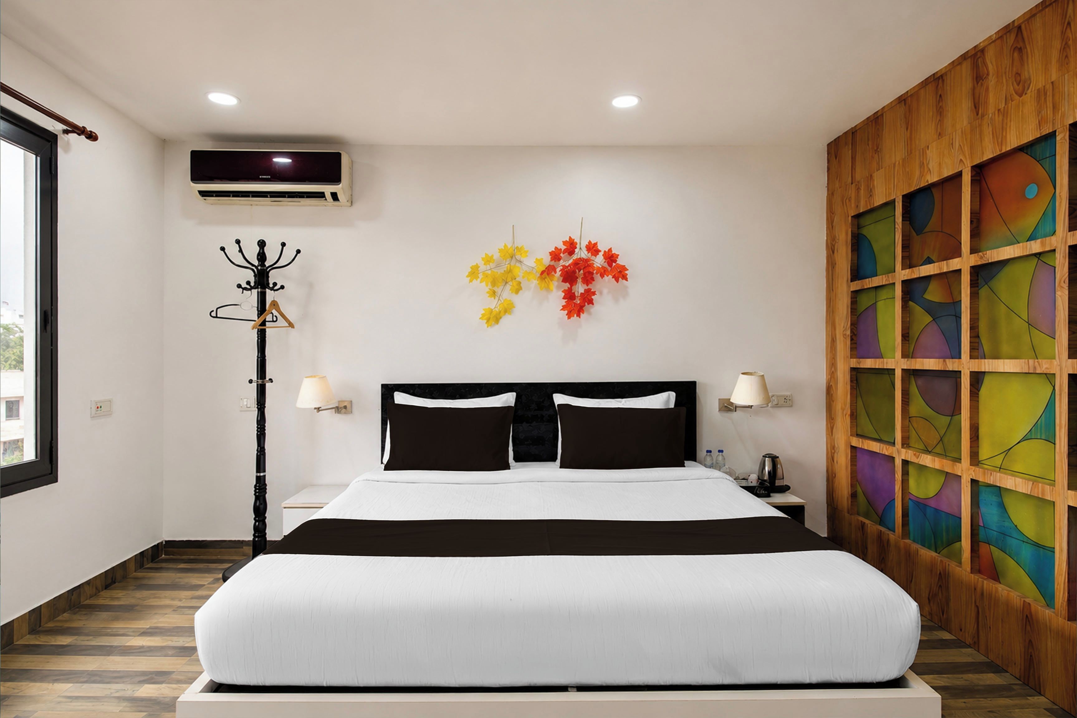 Una camera d'albergo con un letto, un appendiabiti, una lampada e una parete con un motivo astratto dai colori vivaci.