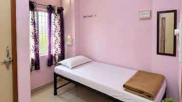 A single-bed room with a ceiling fan, a bedside table with a plant, a window with purple curtains, and a wall-mounted light fixture.