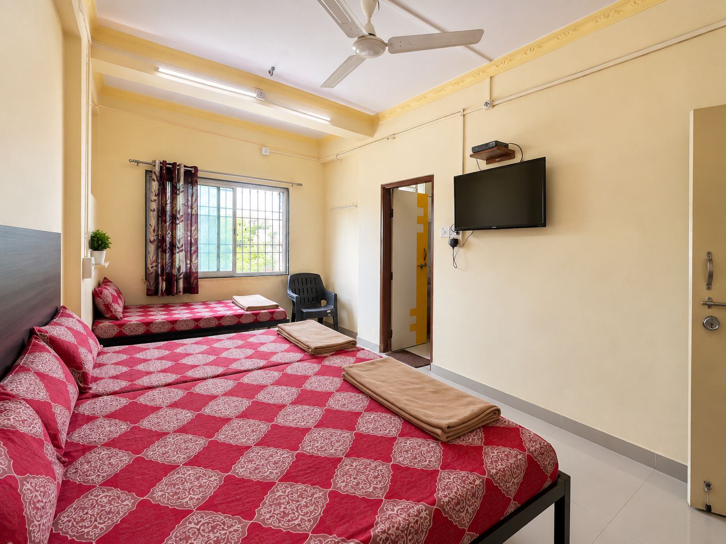 Habitación de hotel con dos camas, televisión, ventilador de techo, ventana con cortinas y una puerta.