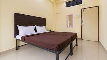 A single bed with a dark headboard and white pillows, a brown bedspread, a small bedside table with a bottle, a mirror with a pink frame, and a door with a handle.