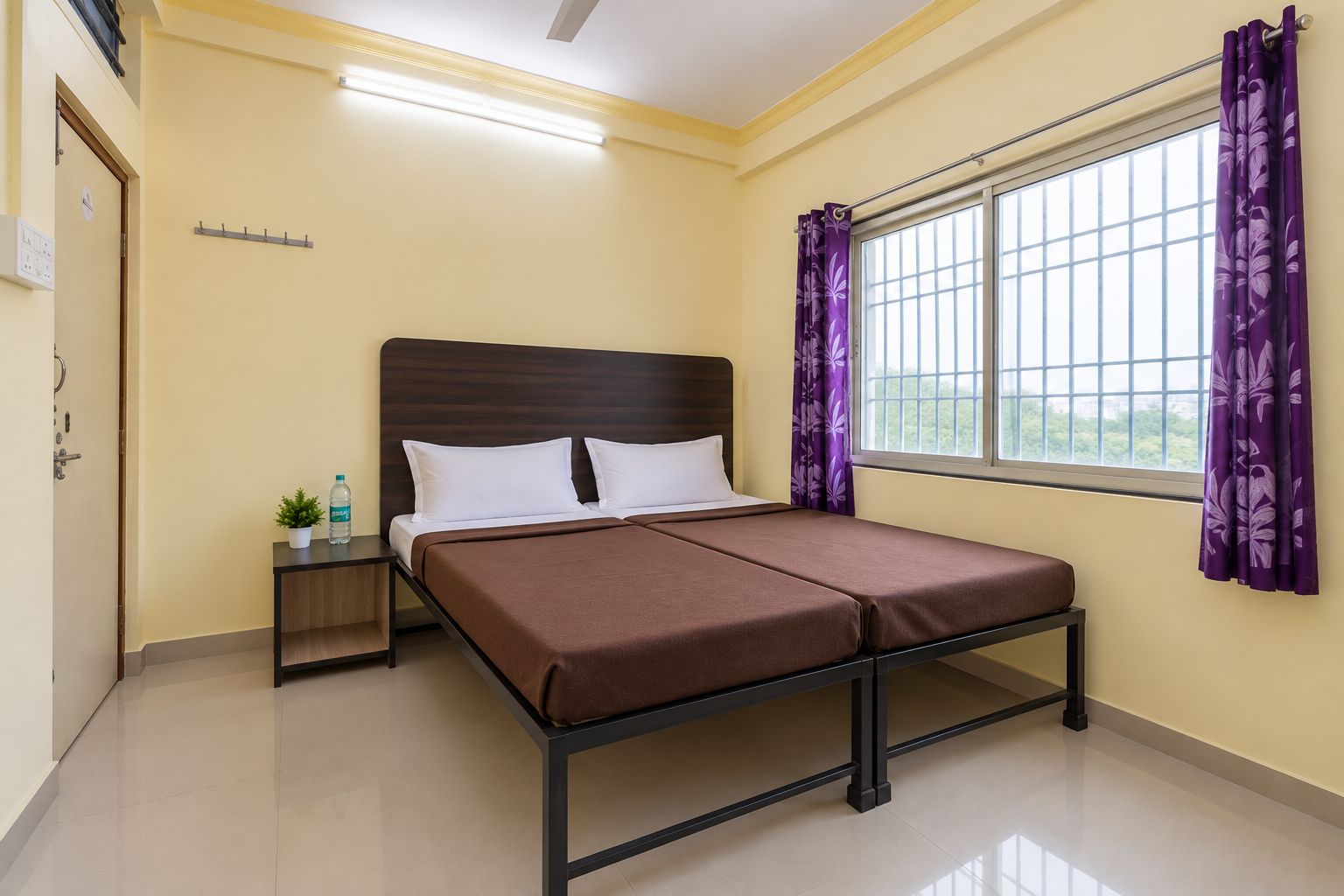 Habitación de hotel con cama, mesita de noche, ventana con cortinas moradas y una lámpara en el techo.