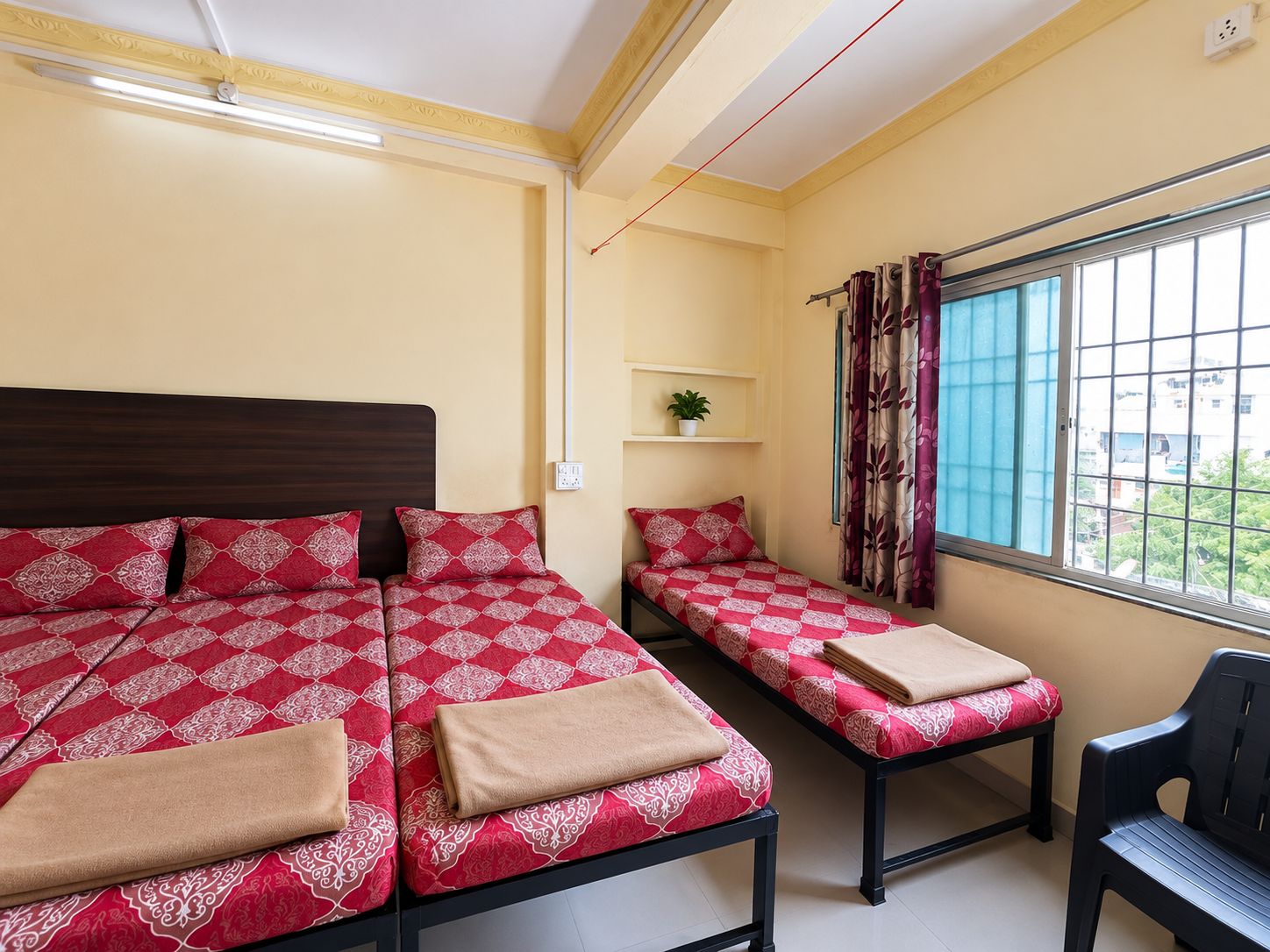 A room with two beds, a chair, a window with curtains, a plant on a shelf, and a power outlet.