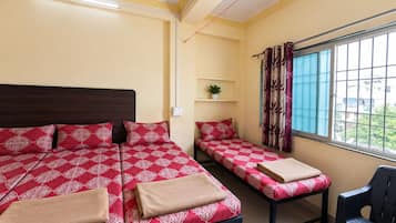A room with two beds, a chair, a window with curtains, a plant on a shelf, and a power outlet.