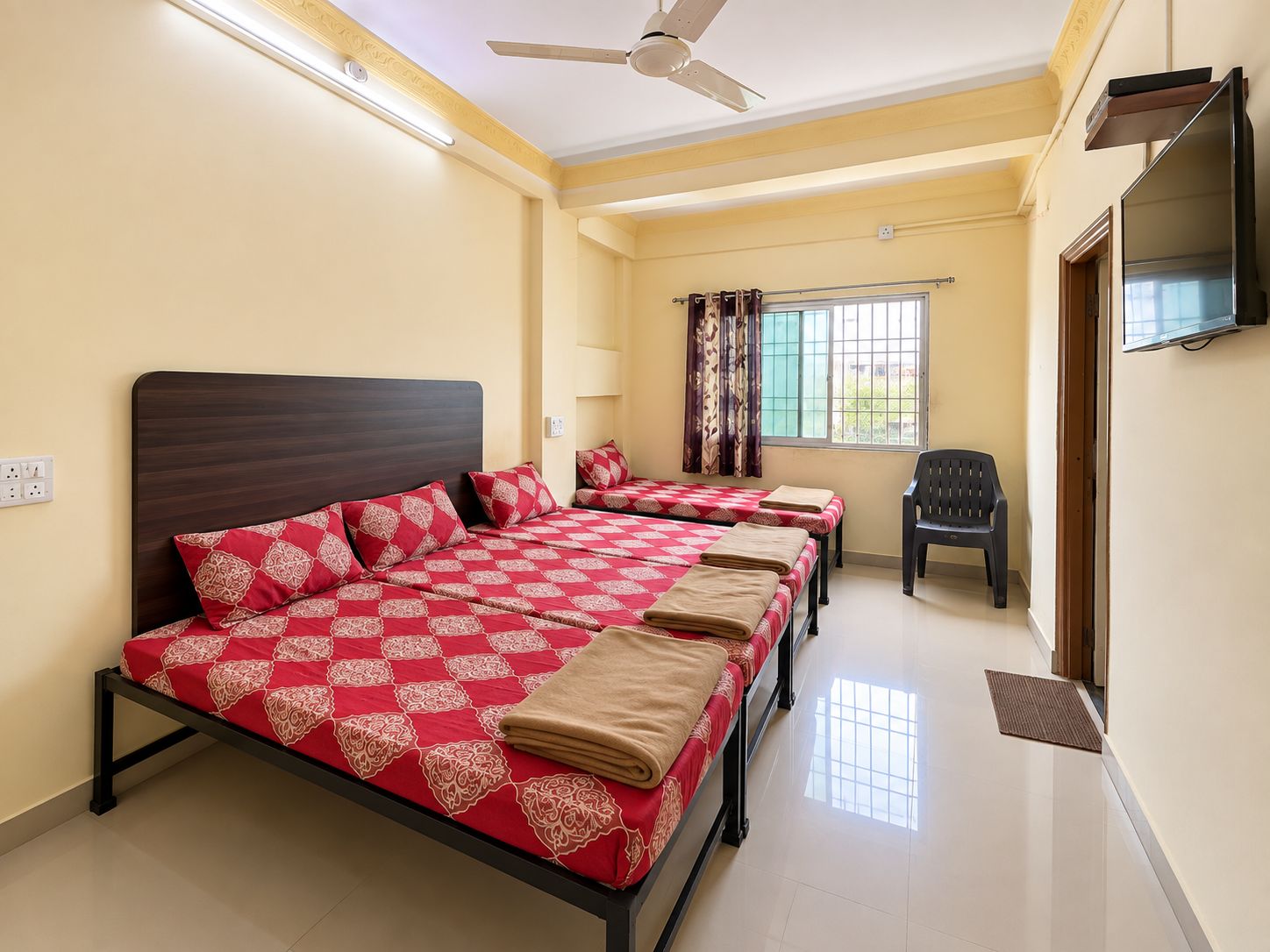A hotel room with a large bed, a smaller bed, a TV, a ceiling fan, and a window with curtains.