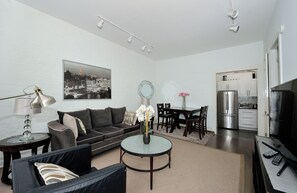 Een moderne woonkamer met een bank, salontafel en een eethoek. Er is een keuken met een koelkast en fornuis.