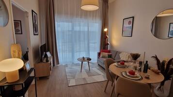 A modern hotel room with a dining area, a sofa, a TV, and a large window with curtains.