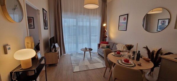 A modern hotel room with a dining area, a sofa, a TV, and a large window with curtains.