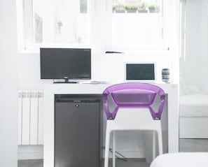 A modern room with a white desk, a black monitor, a white chair, and a window with a view of greenery.