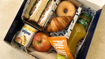 A lunch box containing sandwiches, a croissant, an apple, a juice bottle, a can of yogurt, and a snack bar.