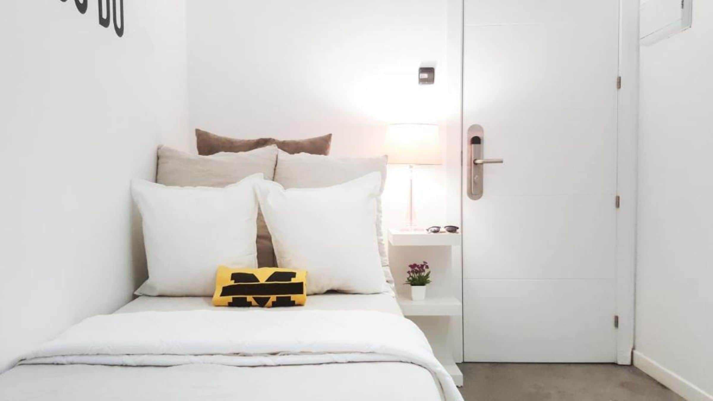 A hotel room with a bed, pillows, a bedside table with a lamp, and a door.