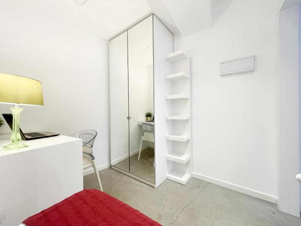 A modern room with a white desk, a laptop, a lamp, a red rug, a closet with a mirror, and a white shelving unit.