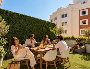 A group of people seated at an outdoor table with a laptop, surrounded by greenery and a building in the background.