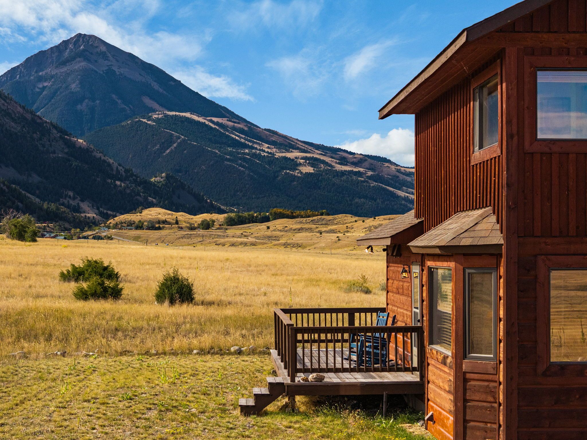 Una cabaña de madera con porche, frente a un paisaje de montañas y campos abiertos.