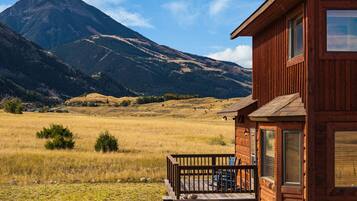 Una cabaña de madera con porche, frente a un paisaje de montañas y campos abiertos.