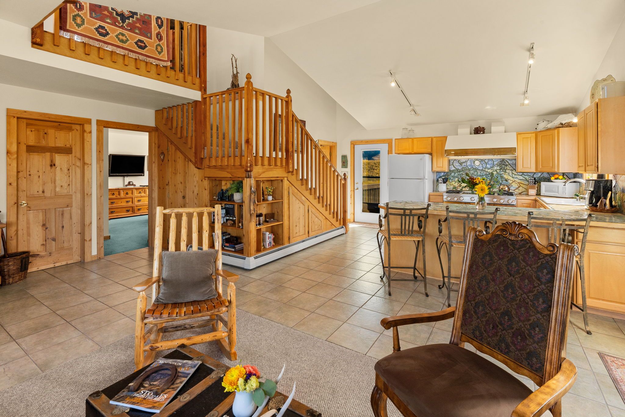 A spacious living area with a wooden staircase, a dining area with a kitchen in the background, and a cozy seating arrangement.