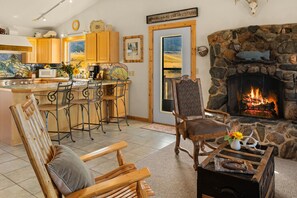 A cozy living room with a stone fireplace, wooden furniture, a kitchen with wooden cabinets, and a view of the mountains.