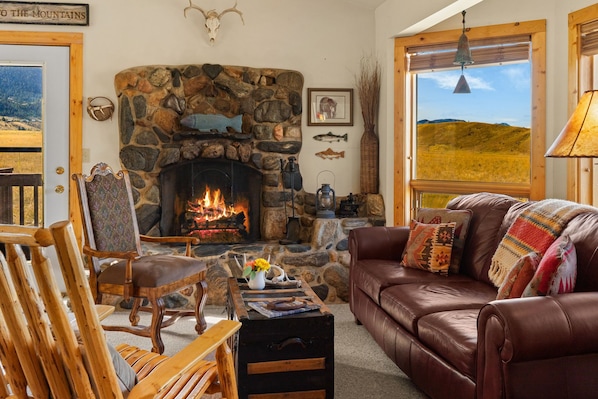 A cozy living room with a stone fireplace, leather sofa, wooden furniture, and a view of the outdoors. - Comfortable, eclectic Montana cabin close to Yellowstone & hot springs (Pray)