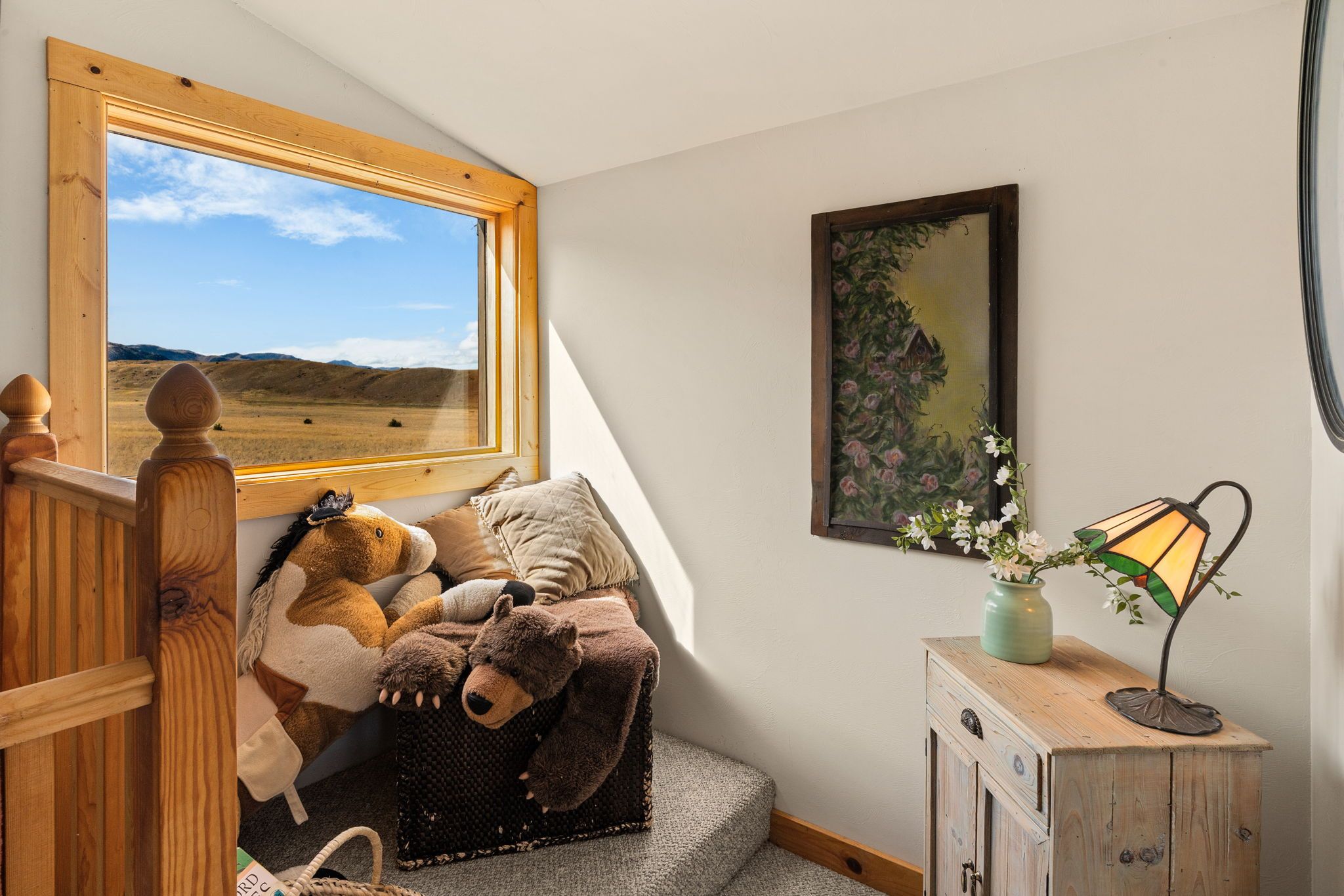 A cozy room with a large window overlooking a landscape, a painting on the wall, and a lamp on a wooden cabinet.