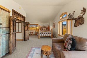 Un intérieur rustique comprenant un lit, un banc en bois, un canapé en cuir et une grande fenêtre donnant sur l’extérieur.