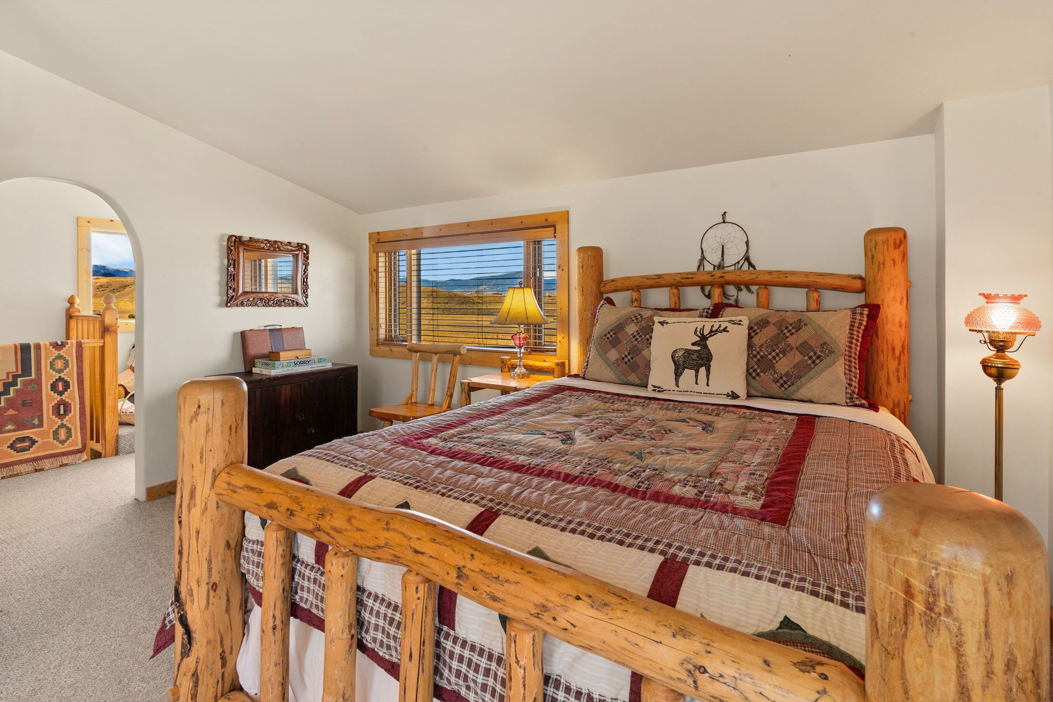 A bedroom with a wooden bed frame, a quilt, a mirror, a lamp, and a window with a view.