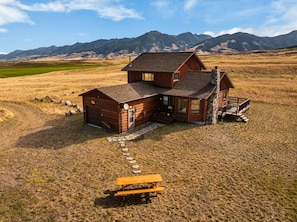 A wooden cabin with a stone chimney, a covered porch, and a picnic table in a grassy field with mountains in the background. - Comfortable, eclectic Montana cabin close to Yellowstone & hot springs (Pray)