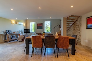 A dining area with a table and chairs, a television, and a staircase with a stone wall. - Livia's House. An 18th-century village farmhouse between Uzès and Nîmes. (Sauzet)