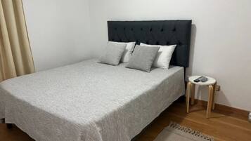 Una cama individual con cubrecama gris y varias almohadas, una mesita redonda con lámpara, y una alfombra pequeña sobre piso de madera.