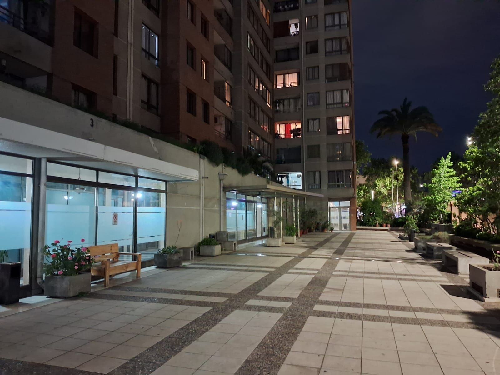 Vista nocturna de un patio de hotel con suelo de baldosas, bancos, plantas en macetas y un edificio con ventanas iluminadas.