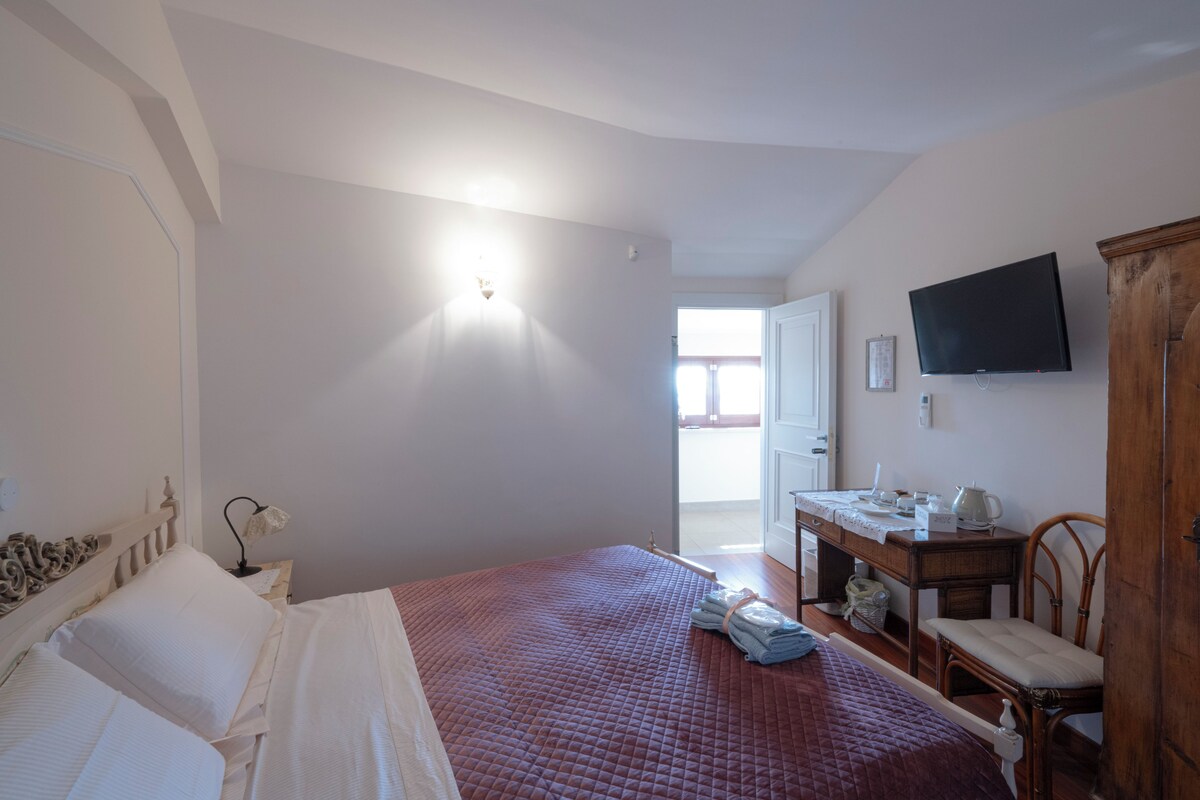 Un dormitorio con cama, escritorio, silla, televisor y puerta que da a un balcón.