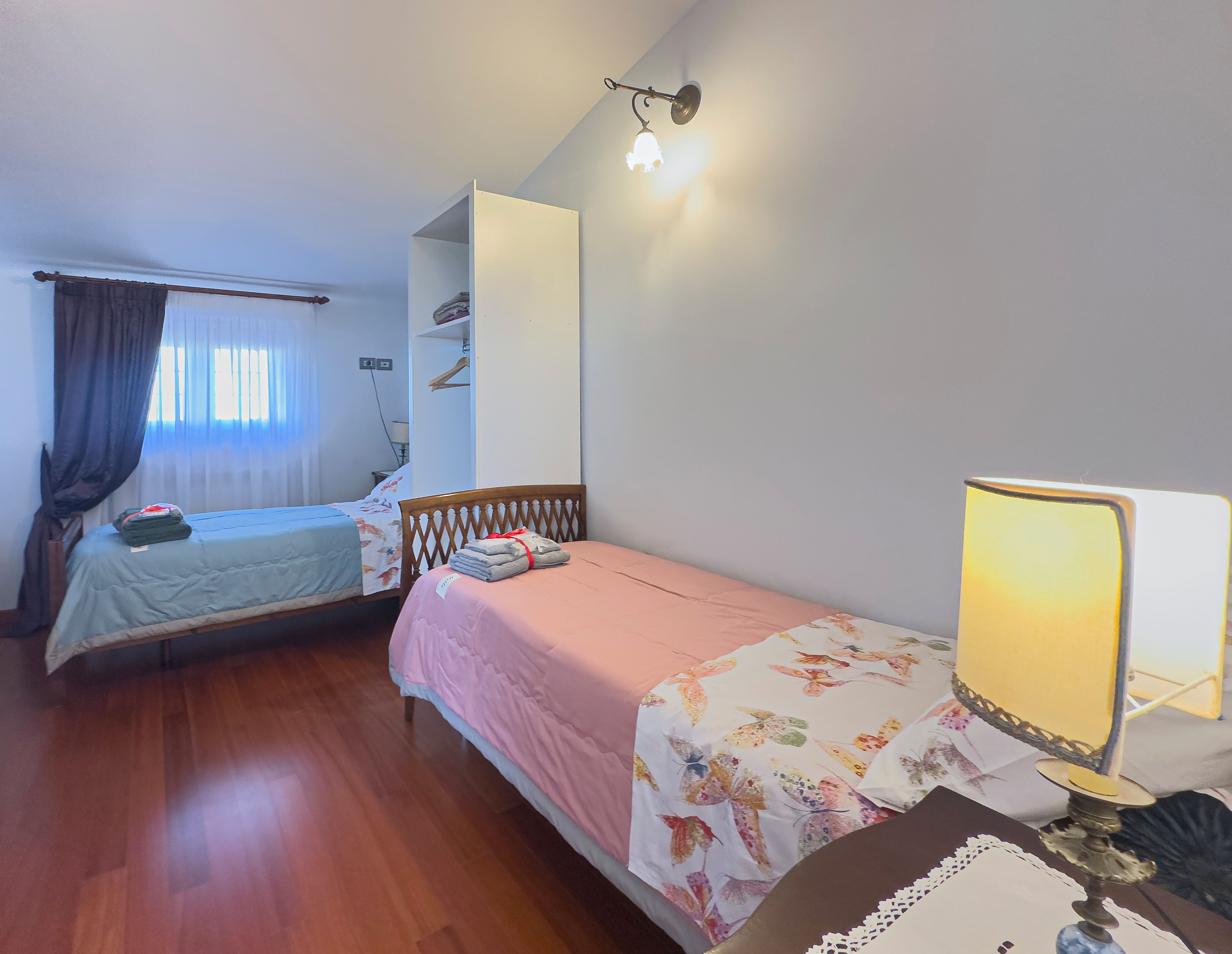 Ein Zimmer mit zwei Einzelbetten, einem Nachttisch mit Lampe, einem Kleiderschrank und einem Fenster mit Vorhängen.