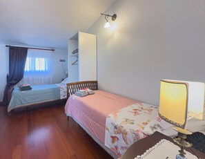 A room with two single beds, a bedside table with a lamp, a wardrobe, and a window with curtains.