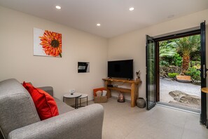 Ein Wohnzimmer mit grauem Sofa, einem roten Kissen, einem kleinen Tisch, einem Flachbildfernseher auf einem Holzständer und einer Tür, die zu einem Garten mit Bank und Palmenbaum führt.