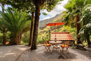 An outdoor seating area with a table and chairs, a red umbrella, and a view of a hillside. - Holiday Home 'Kyra's House' with Wi-Fi (Tabua)