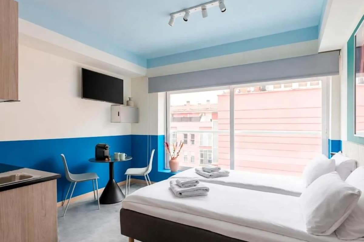 A hotel room with a bed, a desk, a chair, a television, and a window with a view of buildings.