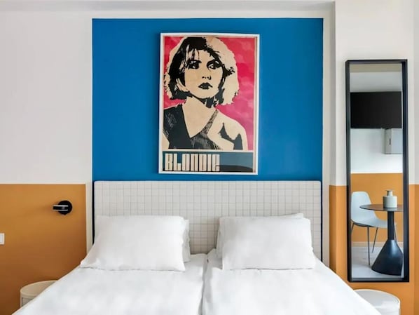 A hotel room with a bed, a wall-mounted artwork, a mirror, and a small table with a chair.