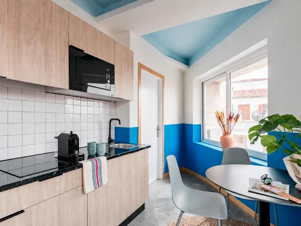 Une cuisine compacte équipée d’un four à micro-ondes, d’une machine à café et d’un petit îlot central. On y trouve également un coin repas avec une table et des chaises, ainsi qu’une fenêtre donnant sur des immeubles.