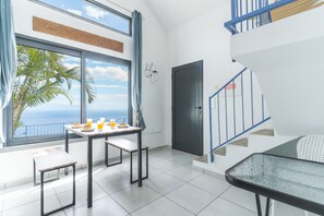 Ein modernes Wohnzimmer mit großem Fenster mit Meerblick, einem Essbereich mit Frühstücksutensilien, einer Treppe mit blauen Geländern und einer Tür mit einem Warnschild für elektrische Geräte.