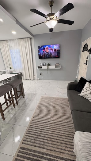 Una sala moderna con una televisión de pantalla plana, un ventilador de techo, un sofá, una mesa de centro y un comedor con mesa y sillas.