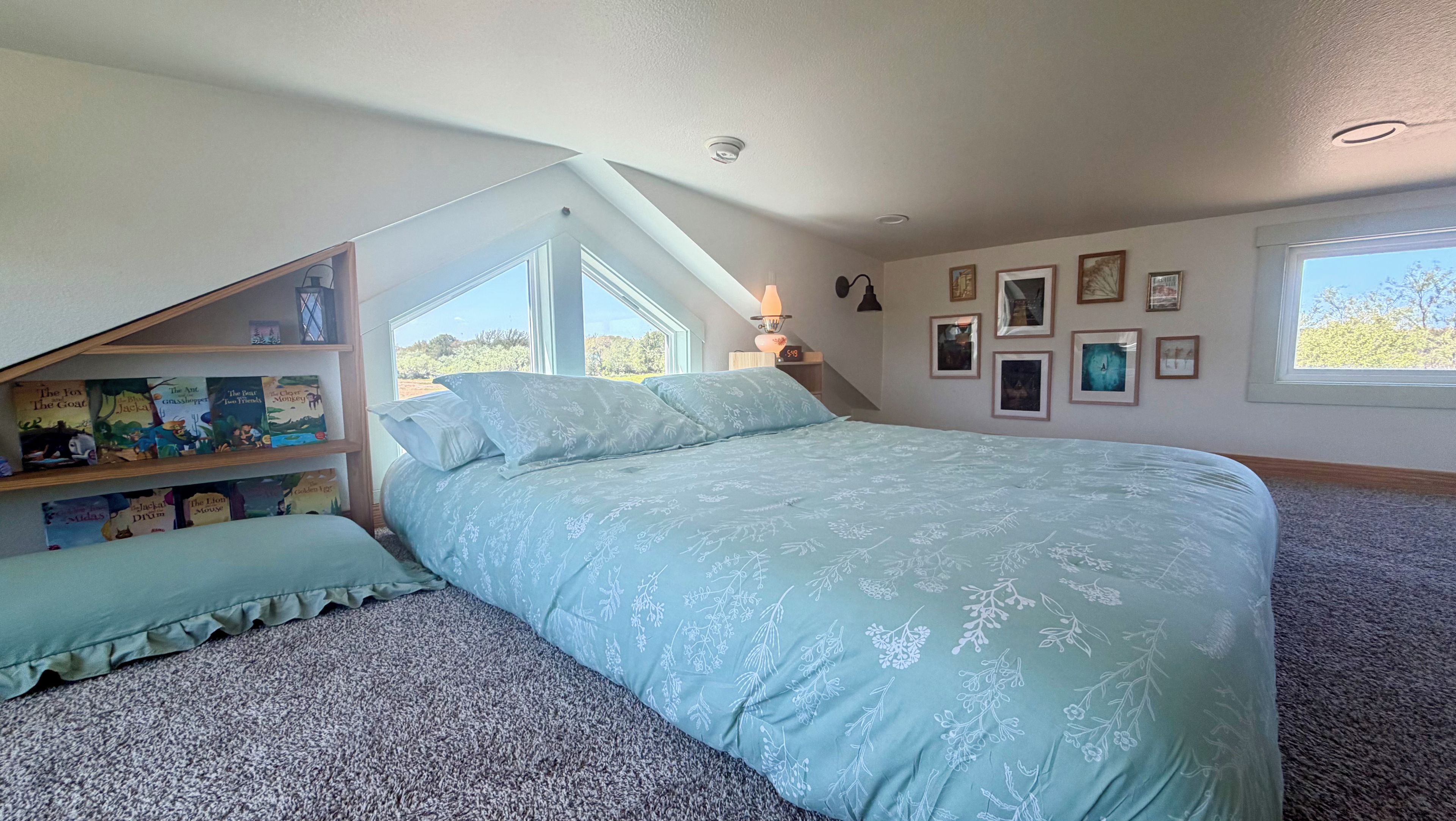 A bedroom with a sloped ceiling, a bed with a floral bedspread, a bookshelf with children's books, and framed pictures on the wall.