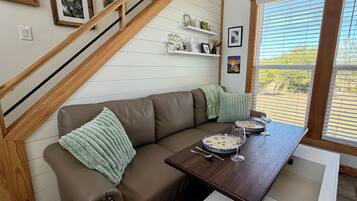 Una sala de estar acogedora con un sofá de cuero marrón, una escalera de madera y una mesa de comedor dispuesta con platos y vasos.