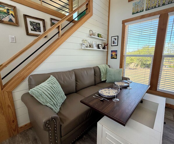 A cozy living room with a brown leather sofa, a wooden staircase, and a dining table set with plates and glasses.