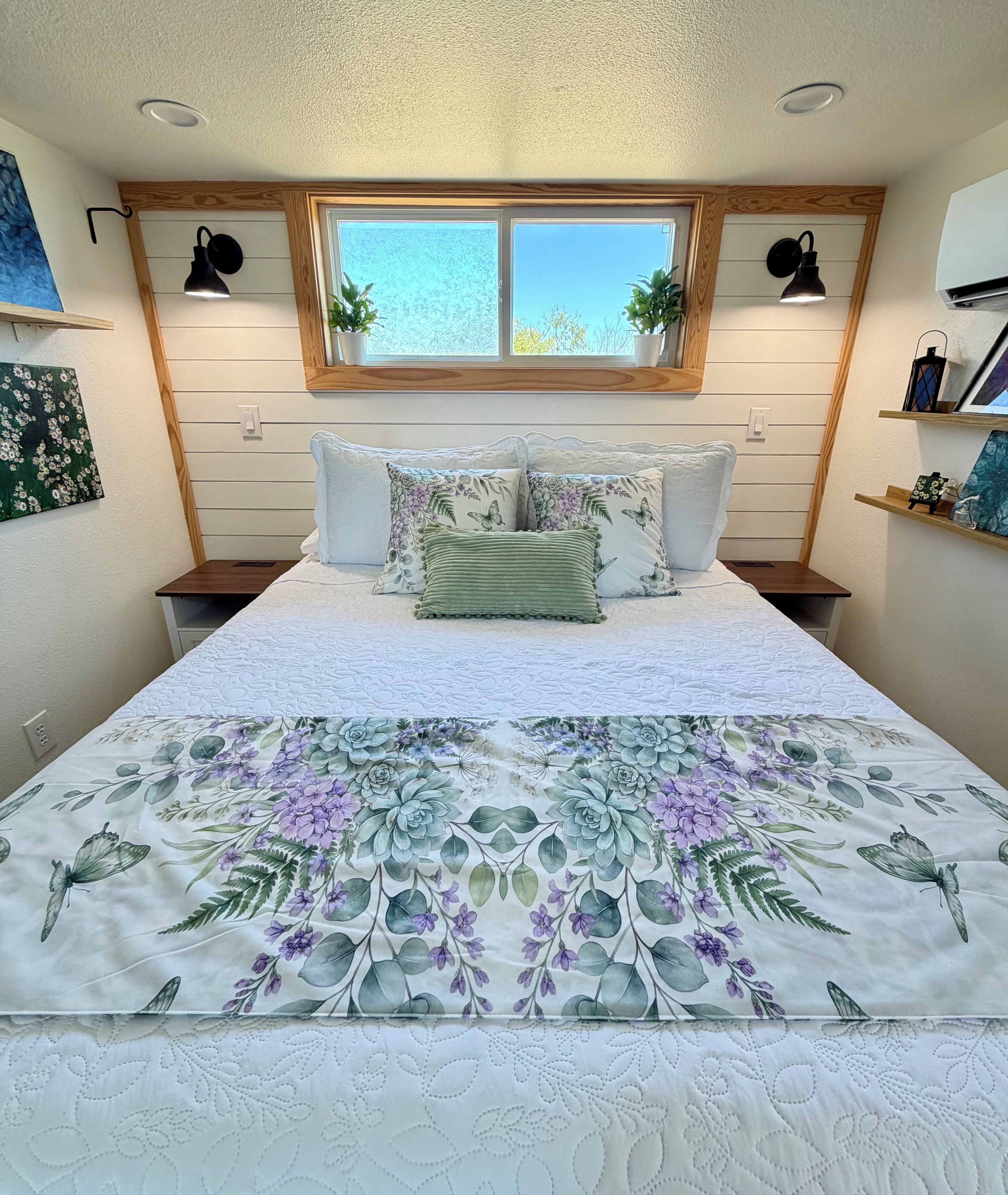 A bedroom with a large bed, a window with plants, and a wall-mounted shelf.