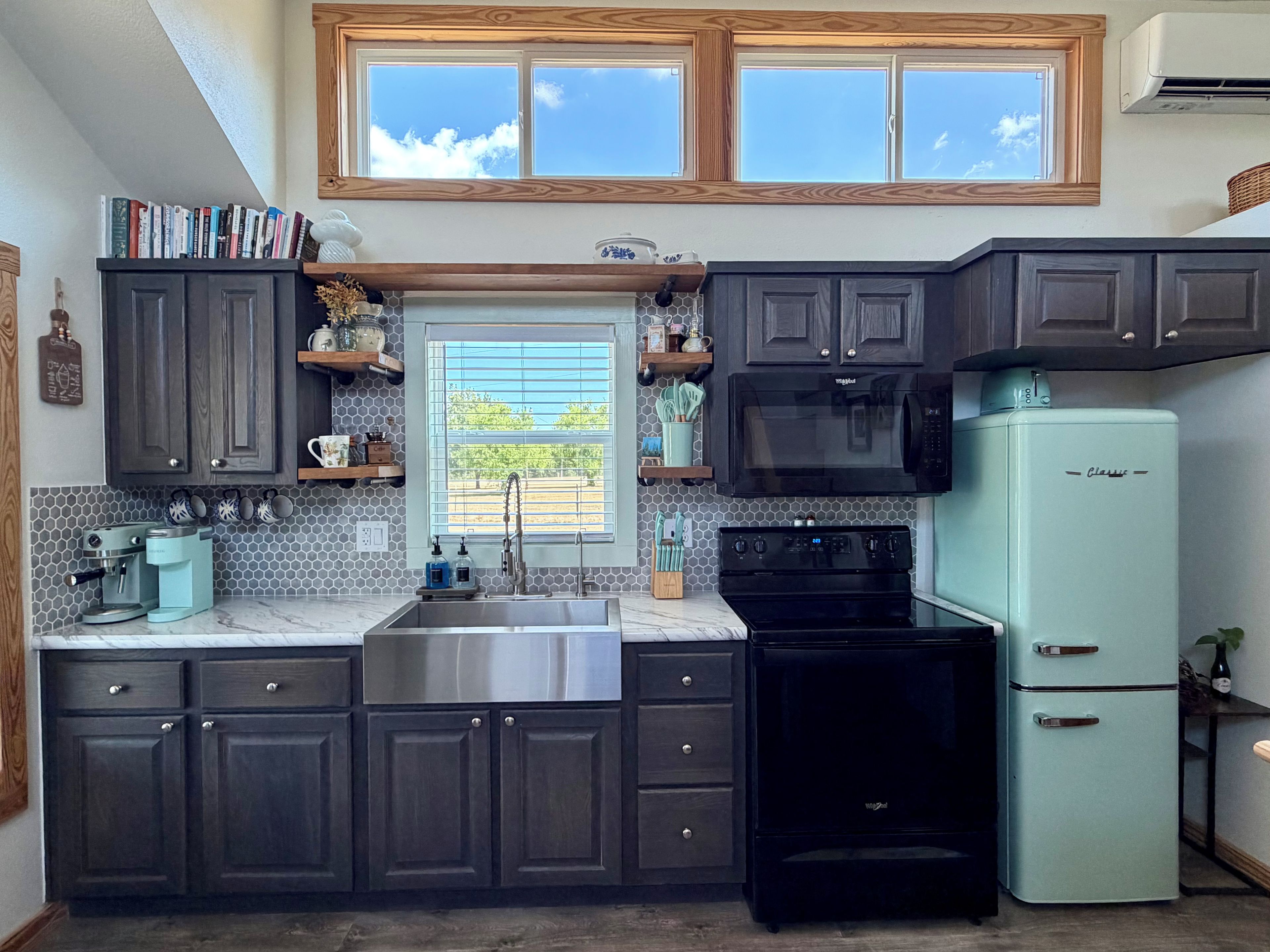 A kitchen with dark cabinets, a stainless steel sink, a black stove, a light blue refrigerator, and a window with blinds.