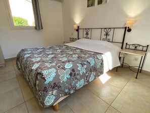 A bed with a floral bedspread, a bedside table with a lamp, and a window with curtains. - Provencal Family Getaway for Six (L'Isle-sur-la-Sorgue)