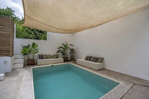 Ein Swimmingpool mit einer weißen Wand und einem schattigen Bereich darüber. Zwei weiße Sofas mit Kissen und Topfpflanzen sind vorhanden.