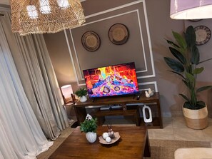 A living room with a television displaying a fireplace, a wooden coffee table with decorative items, a potted plant, and a wall-mounted lamp. - Cozy 2BR Apartment with Tarmac Access (Kampala)