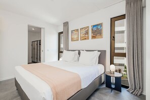 A modern bedroom with a large bed, a bedside table with a lamp, and a view of greenery through a window. - NEW | Harbour Nine at The Wharf | Urban Elegance (Willemstad)